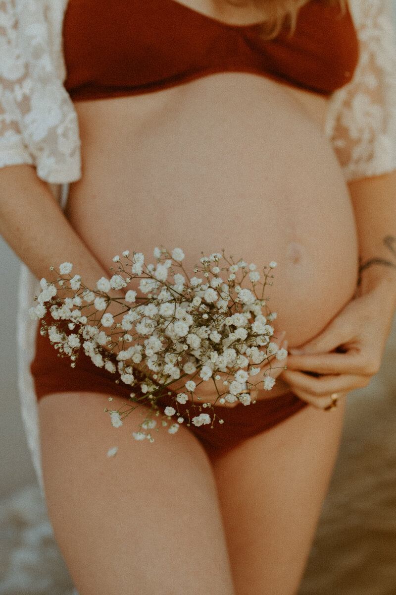 Maternity Photographer, a woman's pregnant belly shows, she wears a deep red swimsuit and holds wildflowers