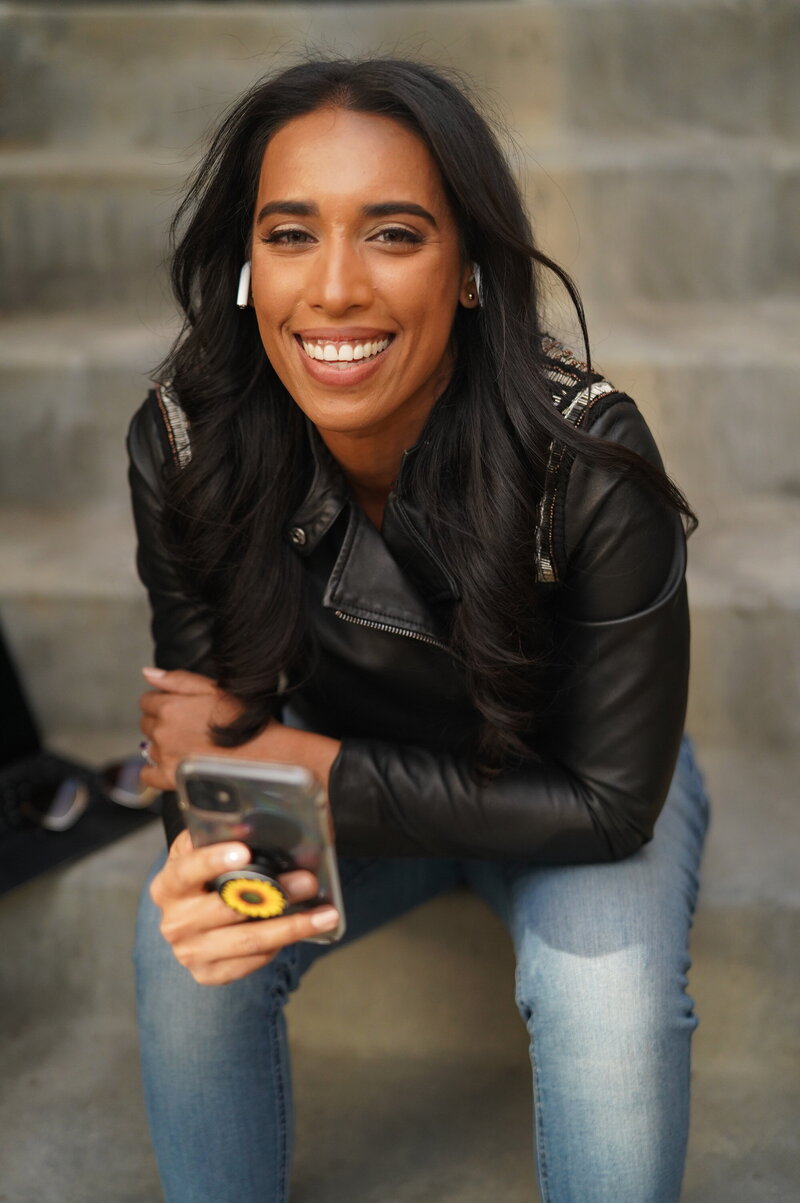 Tania smiling, holding a cell phone, wearing a black jacket and jeans