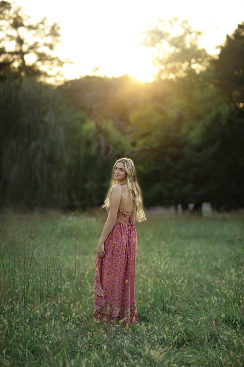 Gorgeous sun-filled senior picture of a pretty girl walking in a field in a floral red dress