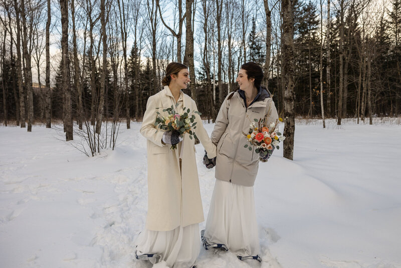 Two brides hold hands and bouquets while walking in the snow on their elopement day.