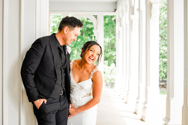 Newlyweds standing on mansion porch laughing together, he is wearing all black and she's laughing off in the distance
