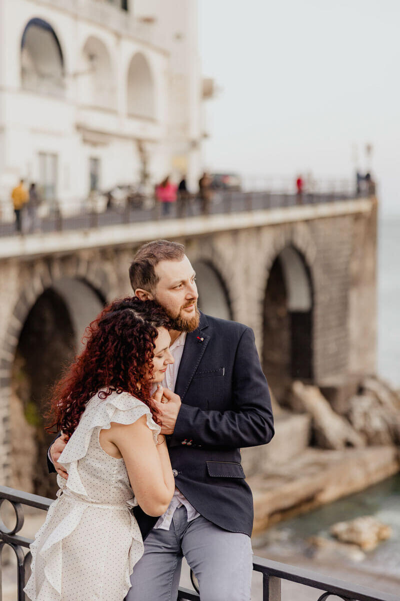 Couple embracing on a balcony overlooking the Amalfi Coast in Italy, captured during a romantic destination engagement photoshoot.