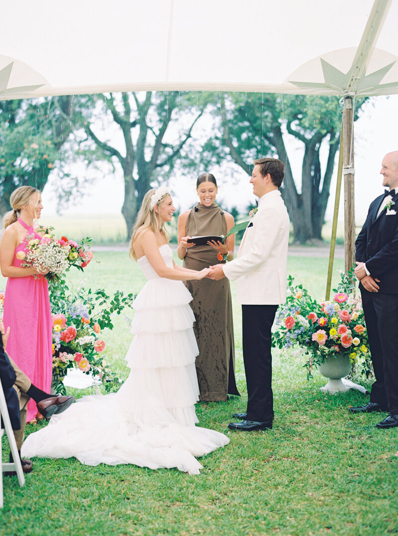 Outdoor wedding ceremony on the lawn at Lowndes Grove. Female wedding officiant in green dress. Bridesmaids in shades of pink. Groom in cream tuxedo jacket. Bride wearing the most gorgeous layered tulle wedding dress and floral themed headband. Charleston destination wedding photographer.