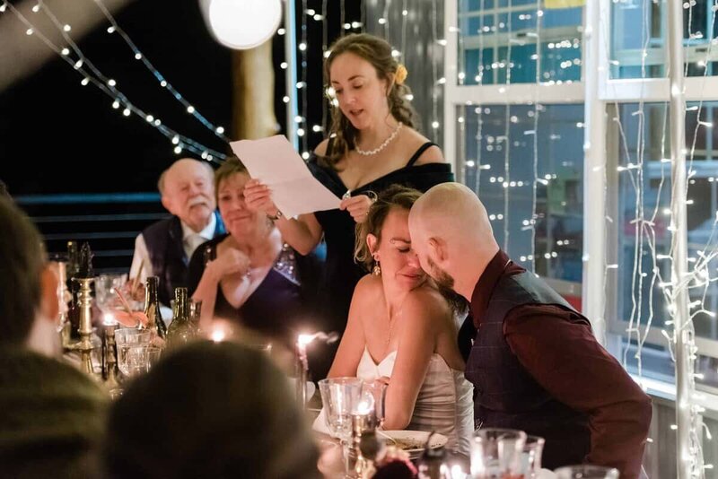 A just married couple leans into each other at a beautifully styled dinner with a long table of family and candles in a twinkle light lit outdoor seating area in the dark.  A family member reads a speech.  Everyone is emotional at this destination elopement wedding in Alaska.