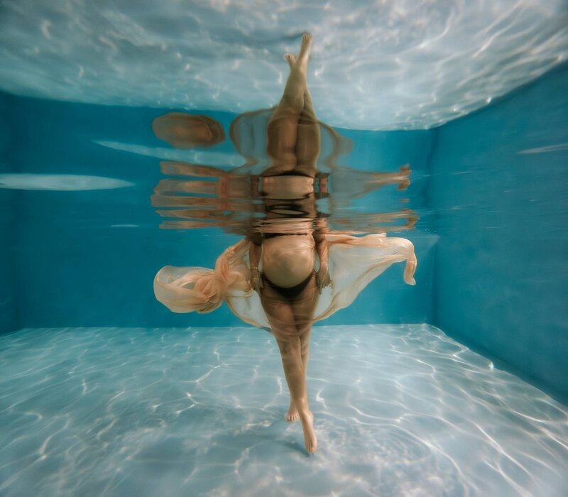 Pregnant woman floating underwater in a maternity photoshoot, with light reflecting off the pool floor as she moves gracefully.