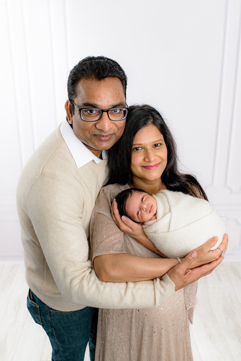 Mother cradles her newborn son in her arms with Father standing behind her wearing glasses and his arm gently supporting her holding the baby