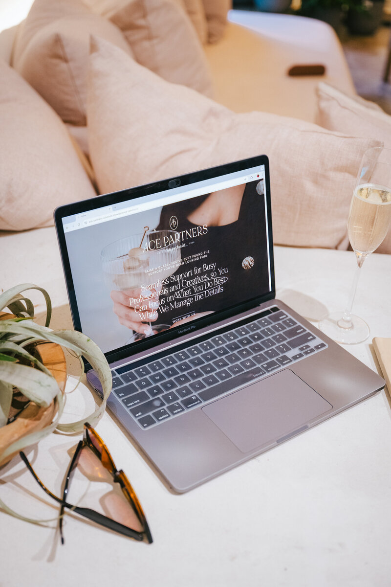 Laptop open to Ace Partners website on white table with glasses, plant, and champagne glass