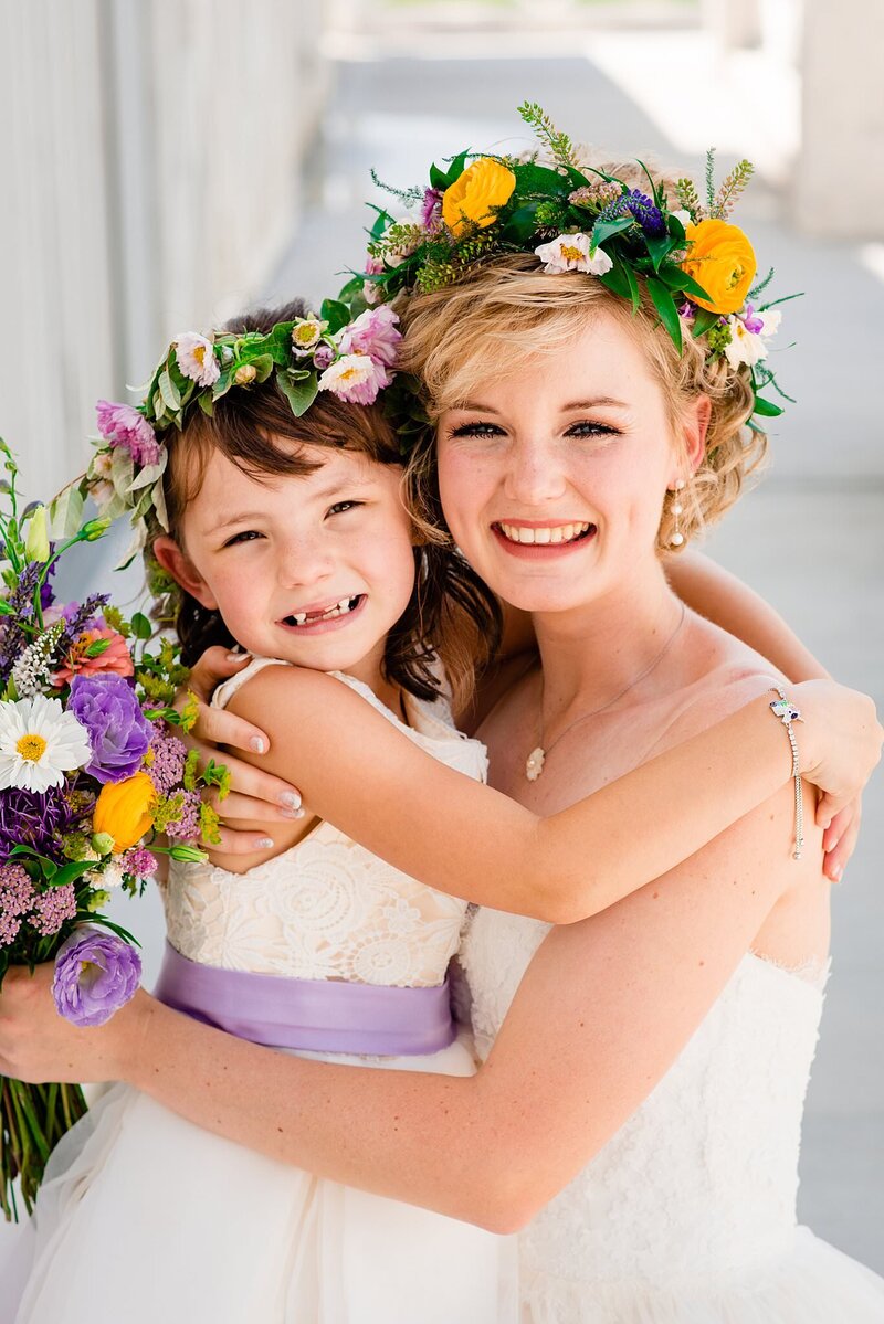Bride hugging flowergirl and smiling at camera, both are wearing bright colorful flower crowns with yellow, pink, and purple flowers