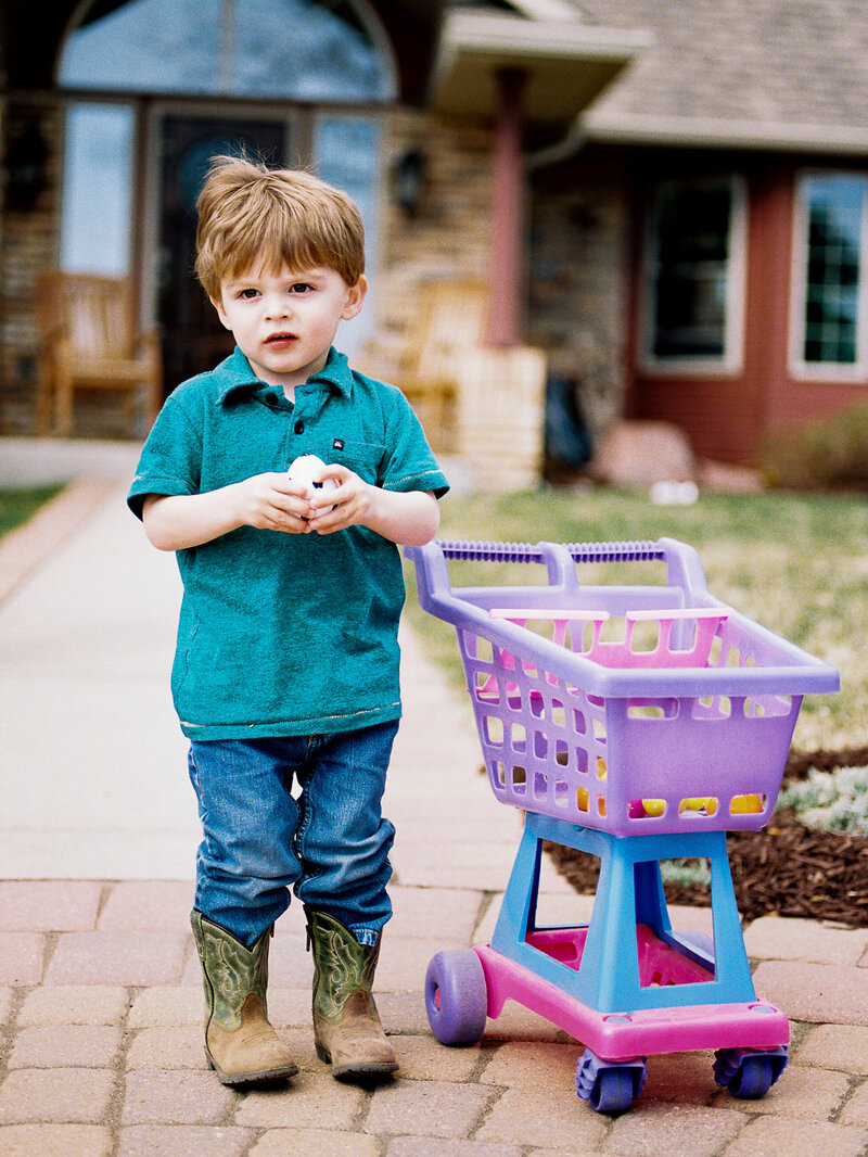 boy in cowboy boots by toy grocery cart in berthoud front yard
