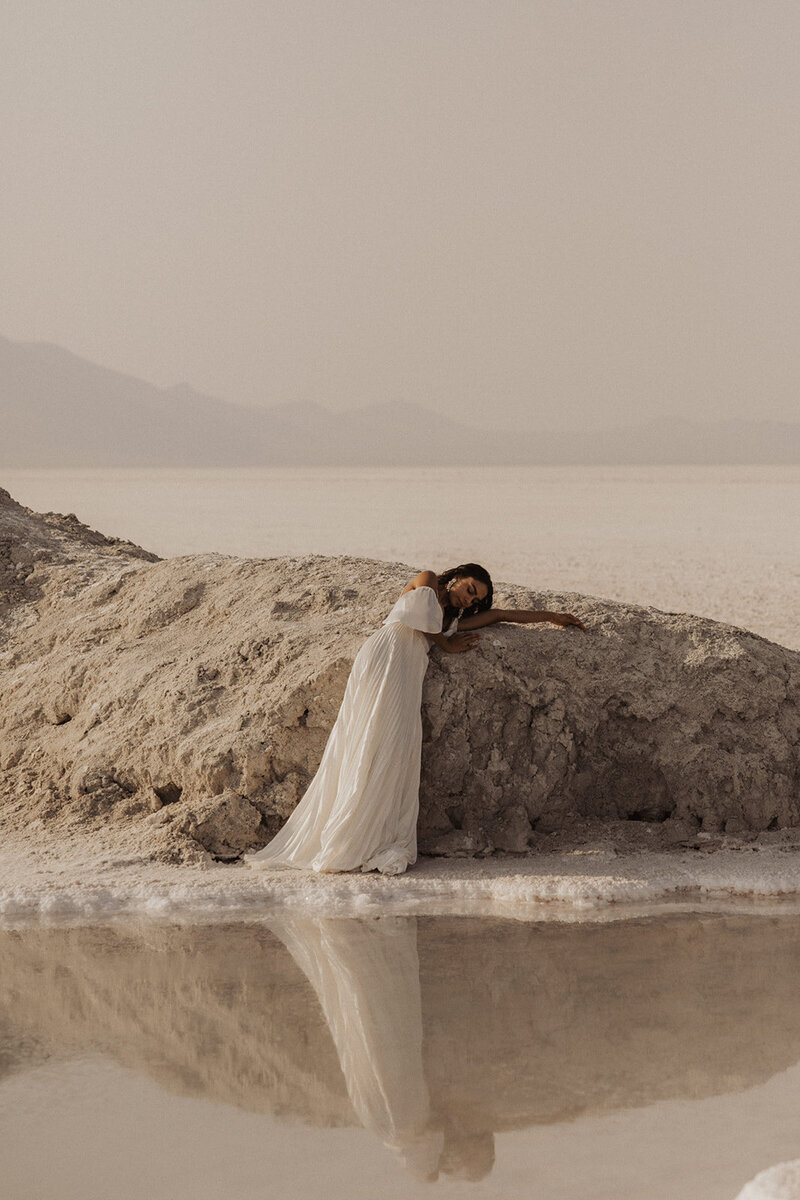 A woman in a white dress leans on a salt rock formation, reflecting in the water.