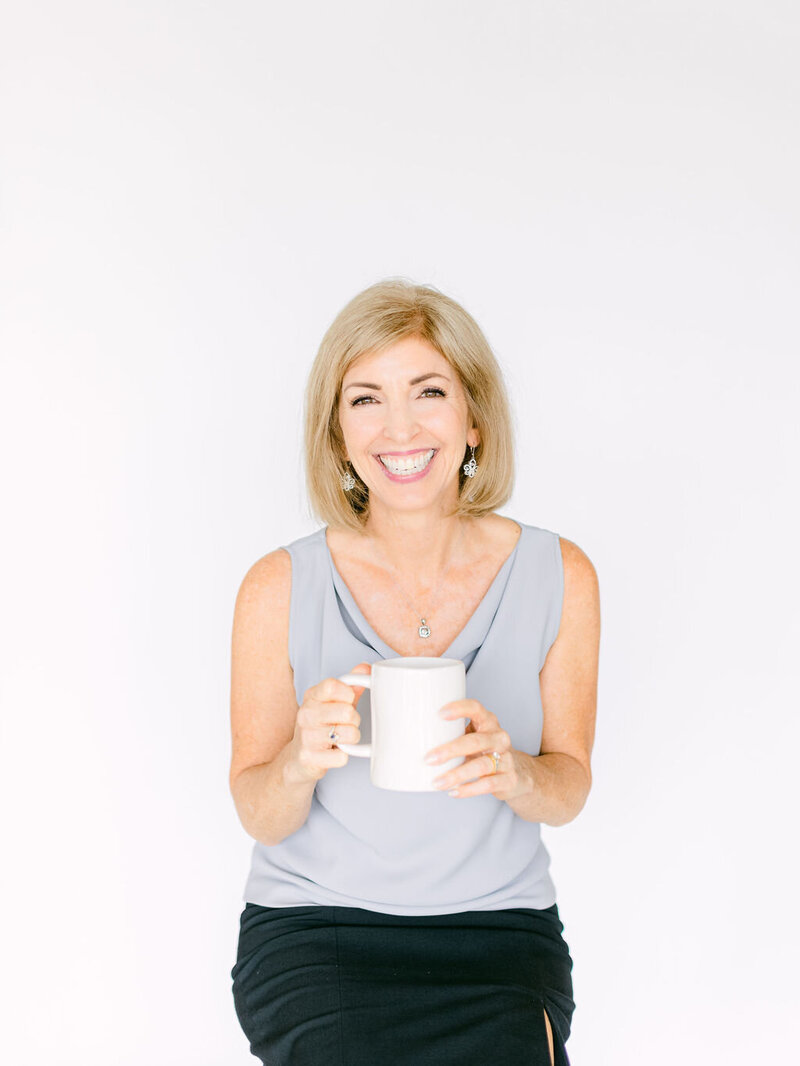 Lora Ulrich in a grey sleeveless top holding a mug, smiling at the camera.