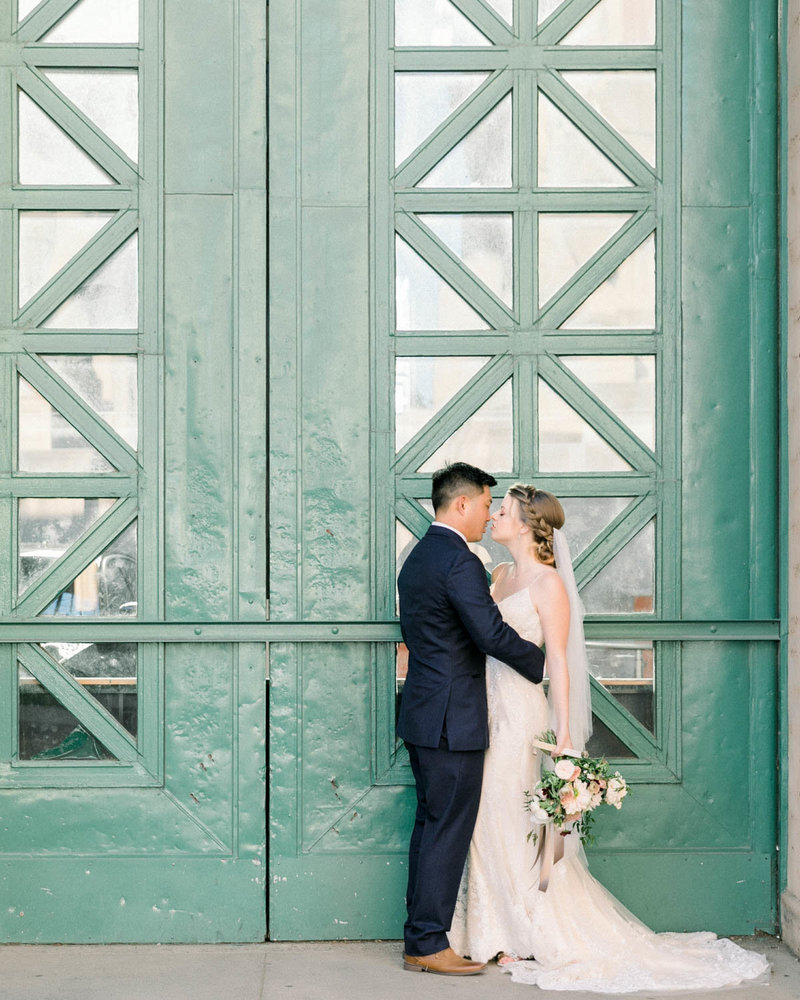 Groom holds Bride as she holds bouquet in front of teal doors
