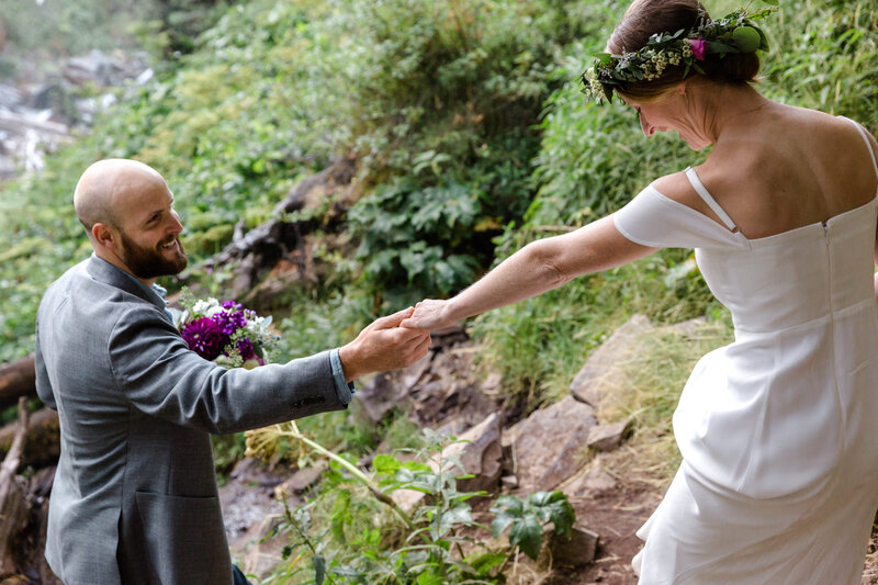 A bald, bearded groom holds a bouquet of purple flowers while reaching up and grabbing his bride's hand as she lifts her white dress up laughing and wearing a floral crown on their Bend Oregon waterfall elopement day at Tumalo Falls. | Erica Swantek Photography