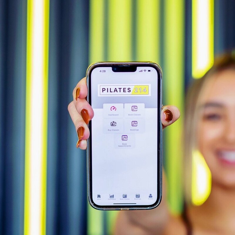 Photo of Pilates instructor holding phone showing the Pilates214 mobile phone app for booking Pilates classes.