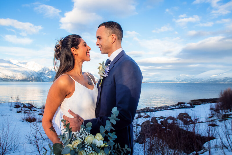 A bride and groom stand together amidst a breathtaking Norwegian landscape on their wedding day. The bride, in a simple and elegant white dress with her hair pulled back, looks lovingly at the groom, who is smartly dressed in a dark navy suit with a tie and boutonniere that complement her bouquet. The bouquet features white and greenery, harmonizing with the natural surroundings. They share a smile, with the serene fjord and snow-dusted mountains behind them under a clear blue sky, creating a picturesque and romantic scene.