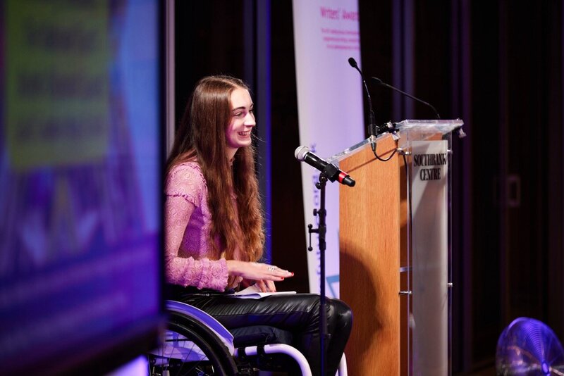 Bethany Handley speaking from her wheelchair on stage at Southbank Centre, smiling beside a microphone and podium during a live event