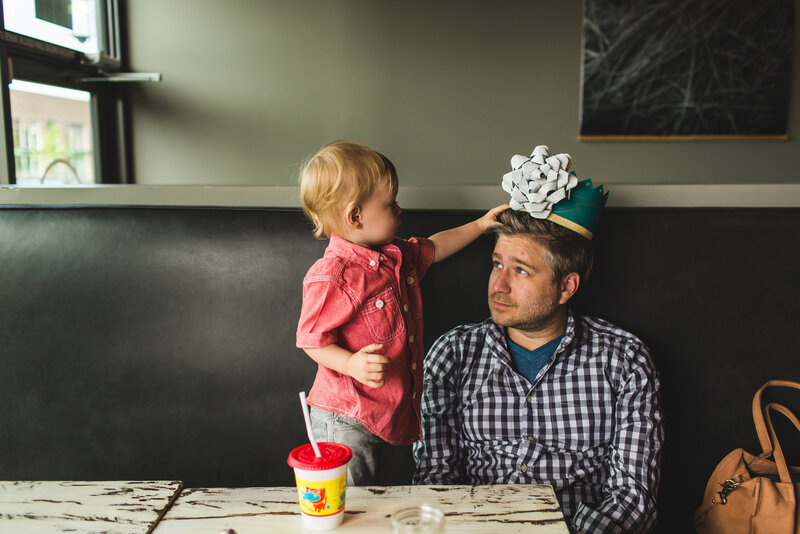 Son putting birthday hat on dad's head at restaurant.