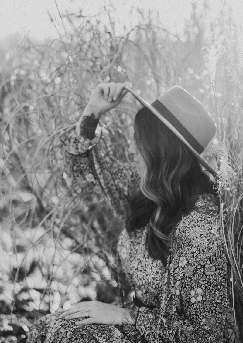 Black and white self portrait of woman holding front of hat  in field of tall grass