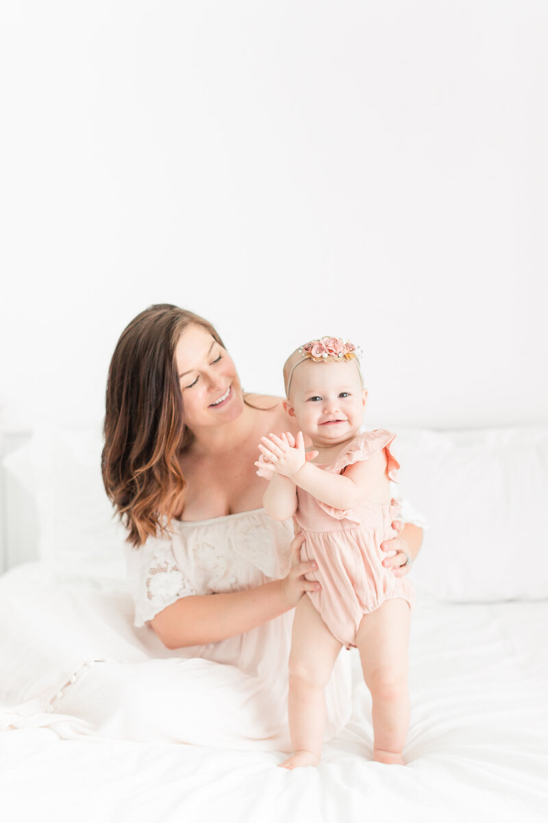 A photo of a mother wearing a maxi white dress holding her daughter in a pink bubble romper smiling at the camera by Washington DC Family PhotographerWashington DC Family Photographer