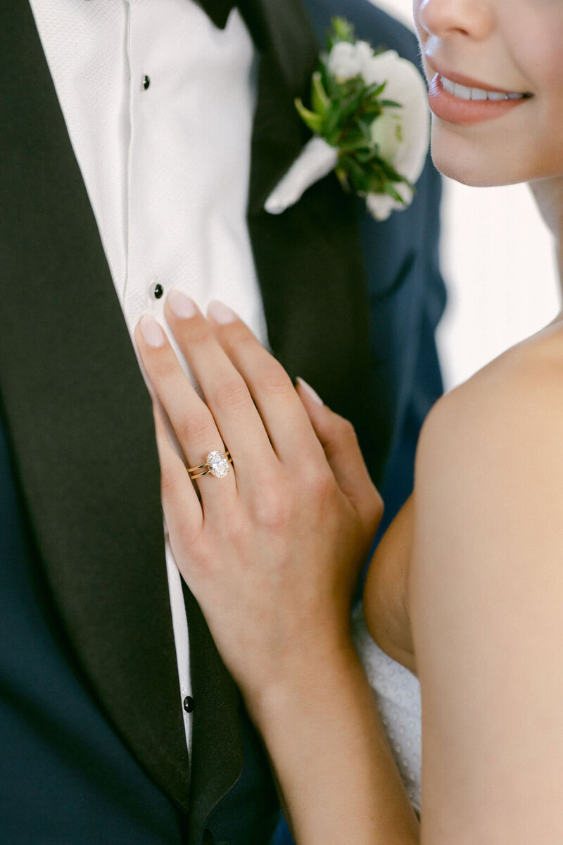 Close up ring shot of bride's oval wedding ring as she delicately places her hand on her groom's lapel