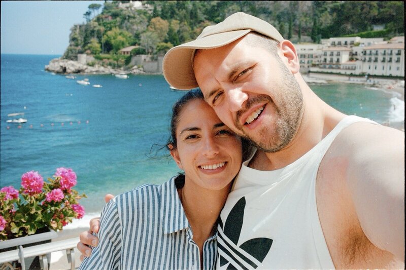 Man and woman pose together with an italian beach behind them
