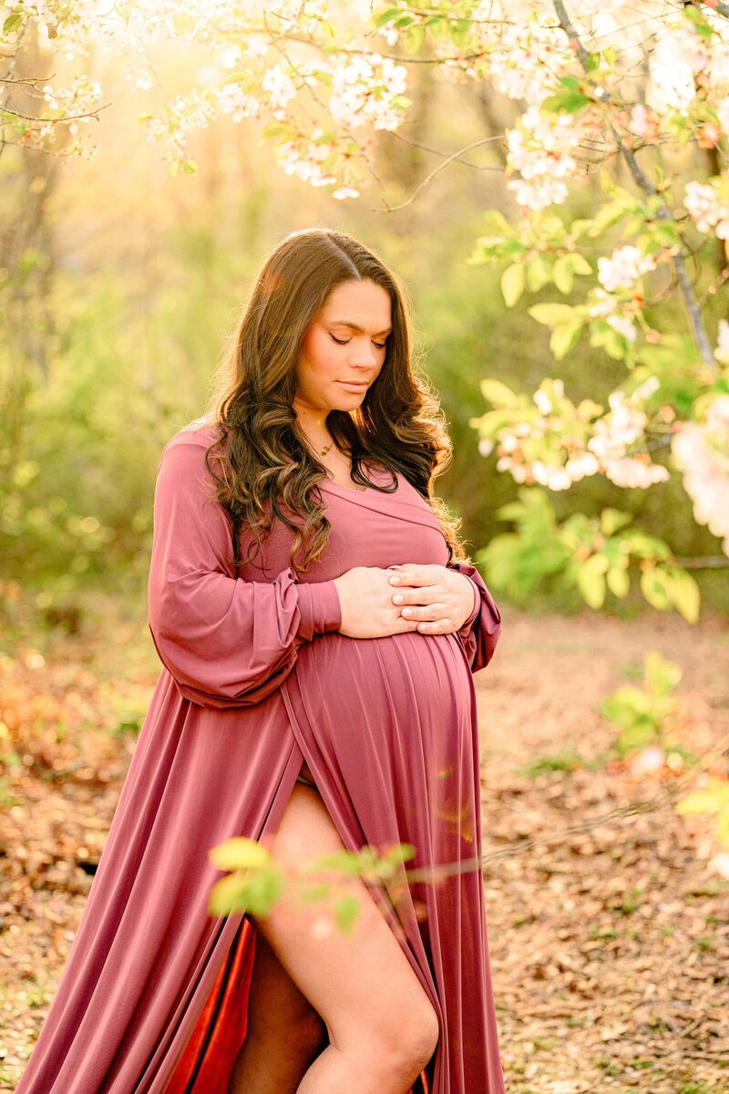 Expectant mother has both hands on the top side of her pregnant belly in a Charlotte Garden area she is dressed and a rose pink dress