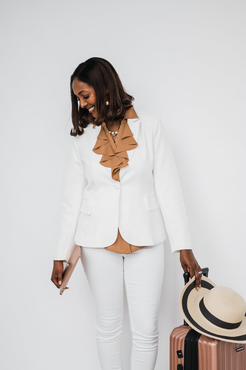 A destination wedding planner in white pants and a blazer holds a suitcase, poised for her next event.