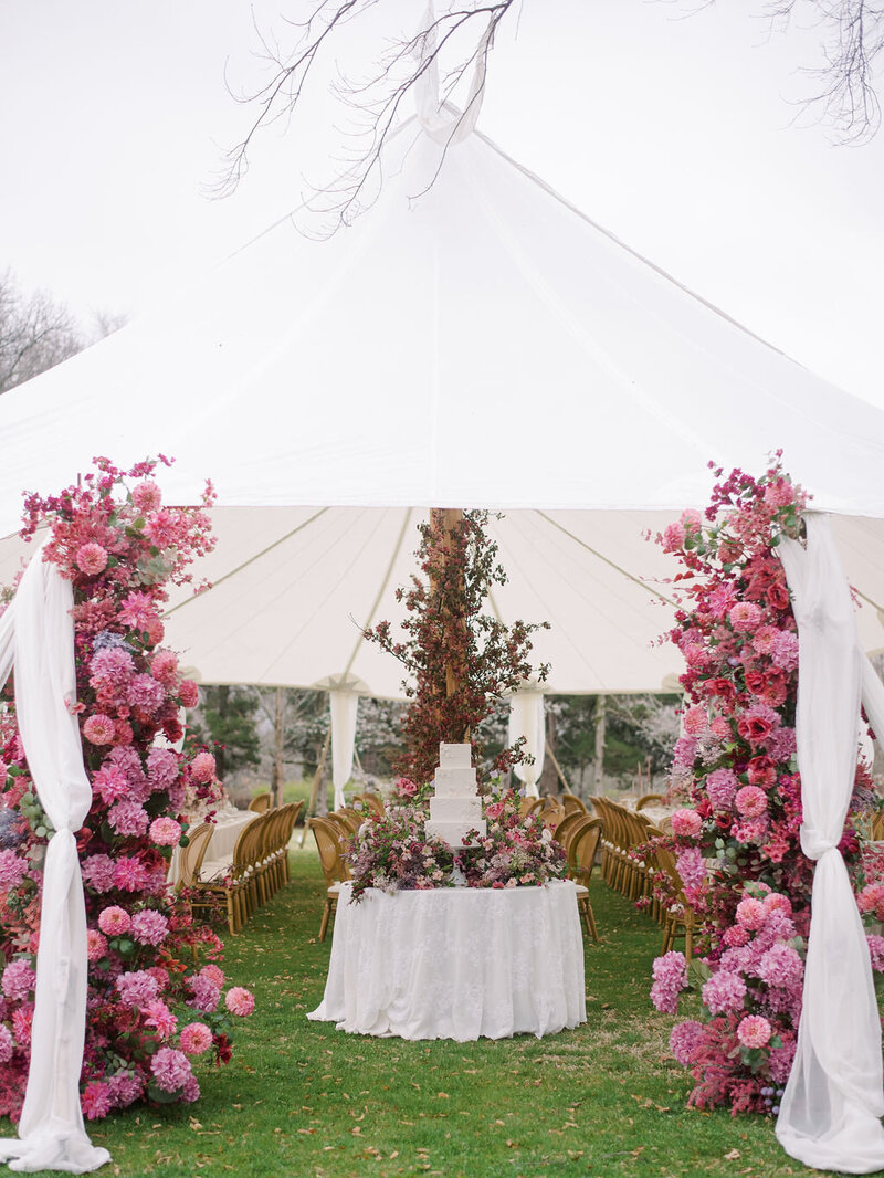 An all-white cake under a reception tent with a magenta floral installation.