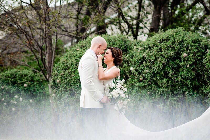 The bride and groom stand in the misty forest and they embrace at Noah Liff Opera Center. The groom is wearing a tuxedo with a white jacket and black pants. The bride is wearing a fitted white dress with a long train. She is holding his cheek while he holds her white floral bouquet.