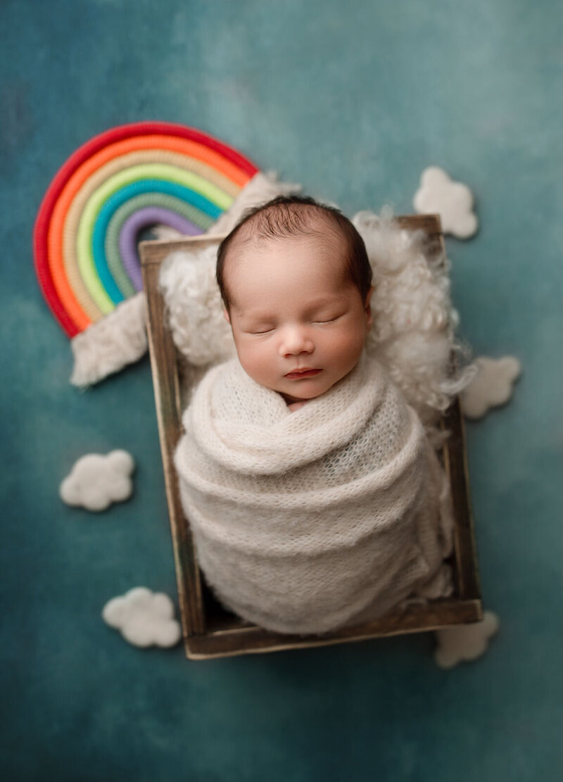 rainbow baby posed in a wooden crate with a rainbow and clouds surrounding it