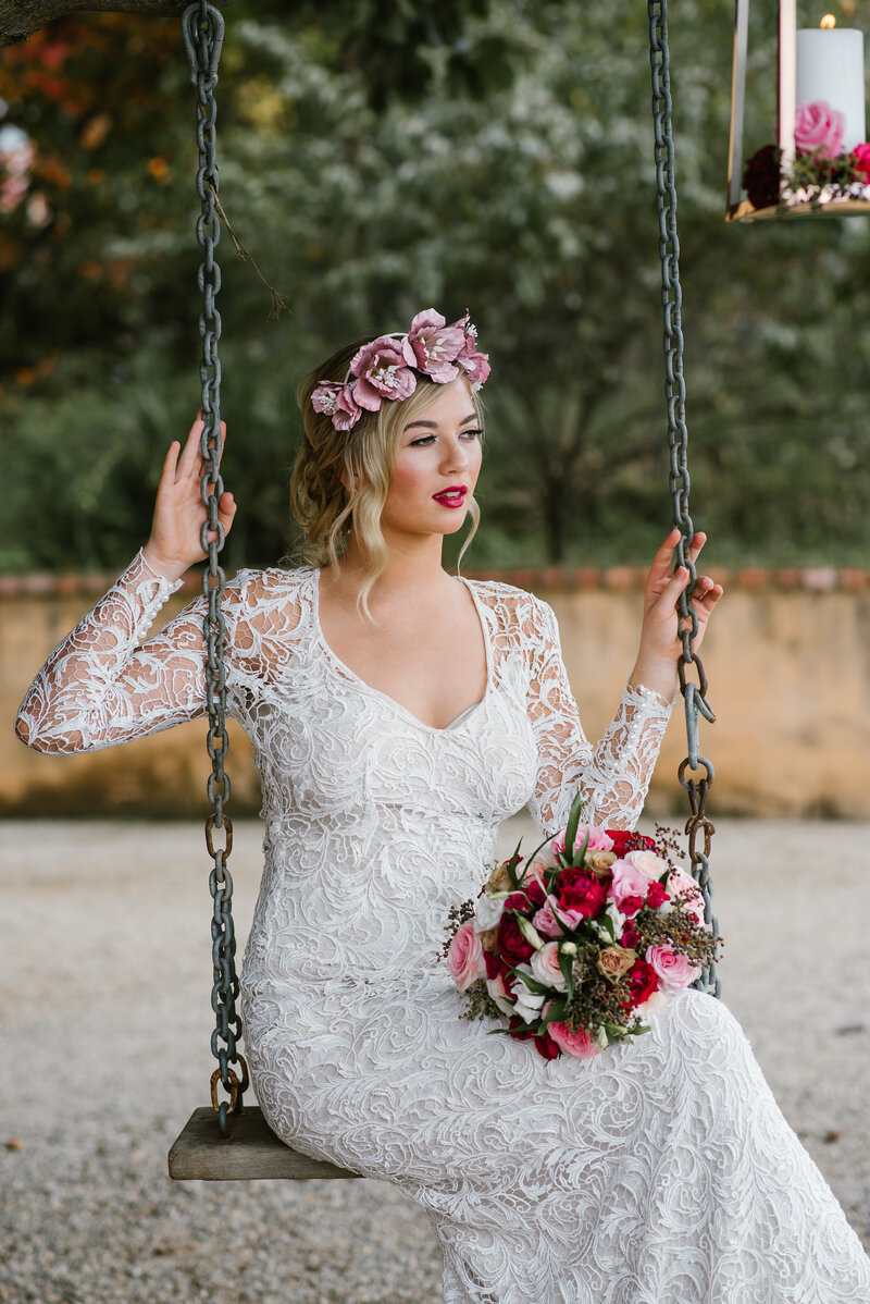 Bride in lace wedding dress sitting on a swing with red and pink bouquet and pink flower crown