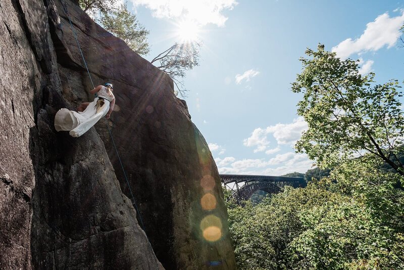 A bride lower down a climbing rope against a dark stone wall with a bridge and blue sky in the distance.  She looks away from her luxury destination elopement photographer, and her long flowing dress blows gracefully in the wind as she lowers down the rope. 