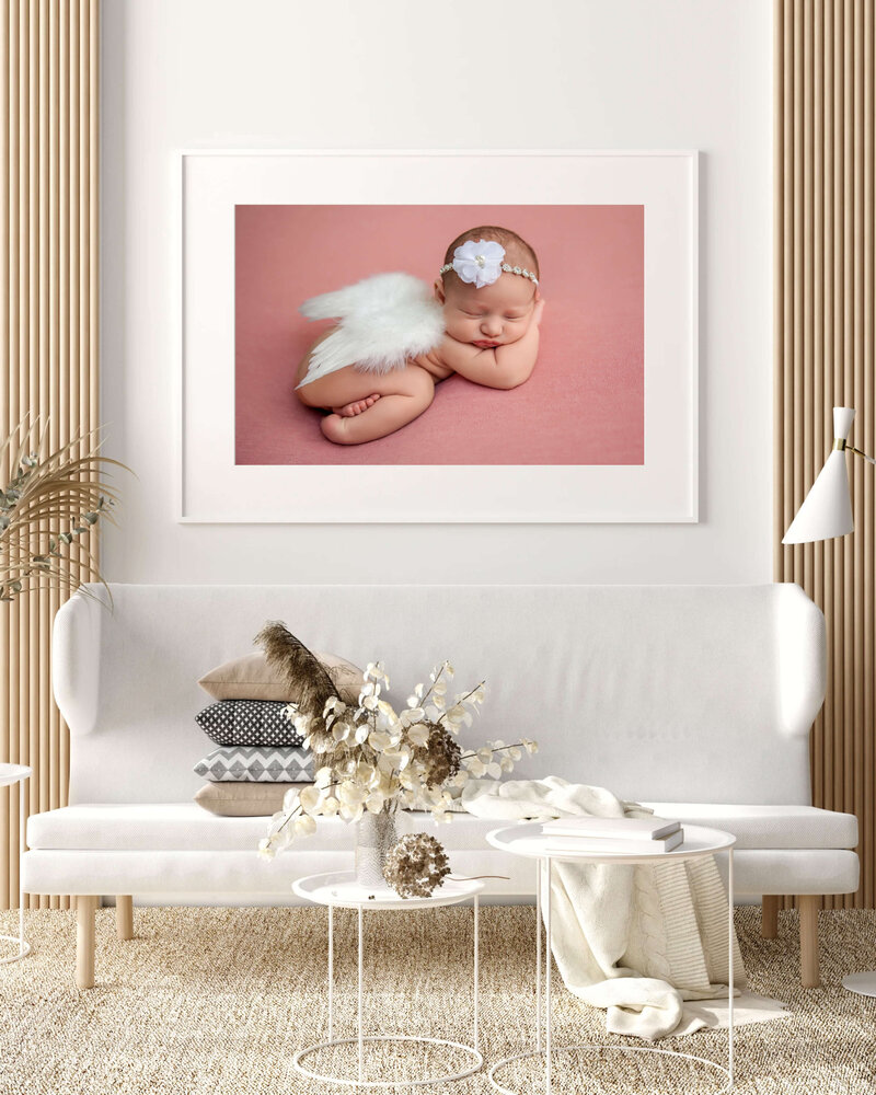 A framed photo by a Jacksonville newborn photographer of a sleeping baby with angel wings and a flower headband hangs above a white sofa in a modern, neutral-toned living room with wooden accents and dried floral decor.