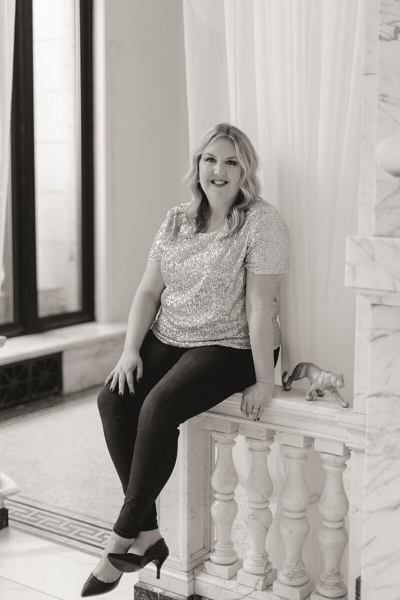 Leadership coach, Melissa Lawrence, smiles in gold sparkly blouse while sitting on marble railing