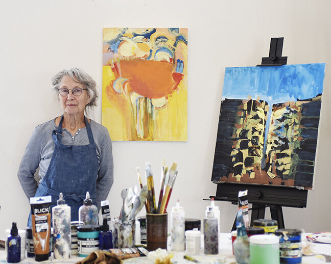 woman standing behind table with art supplies and two paintings