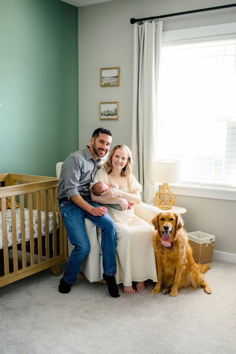 mothe rholds her newborn son in his nursery with dad sitting beside her and brown dog beside them
