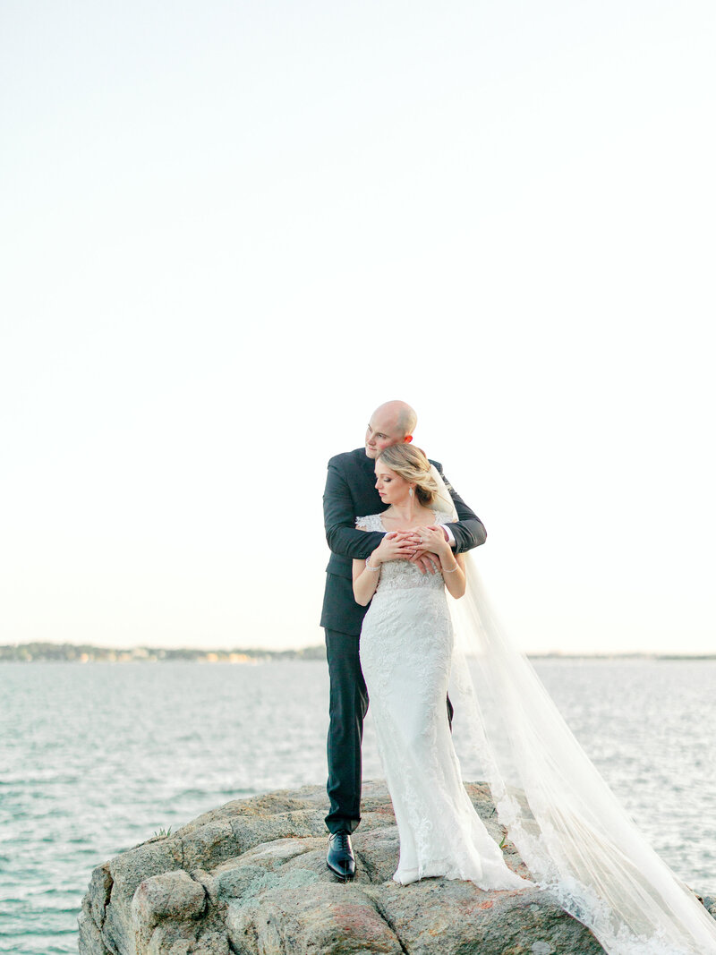 Groom standing behind bride with his arms around her while they stand on a rock in front of the water