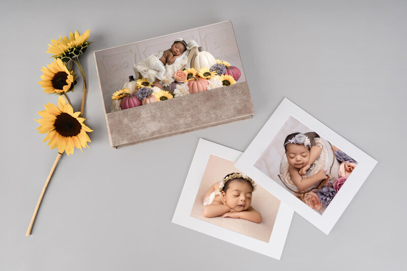 A photo album featuring a newborn baby on the cover is displayed on a gray surface beside two printed baby portraits and a yellow artificial sunflower—perfect for those seeking memorable Newborn photography in Jacksonville.