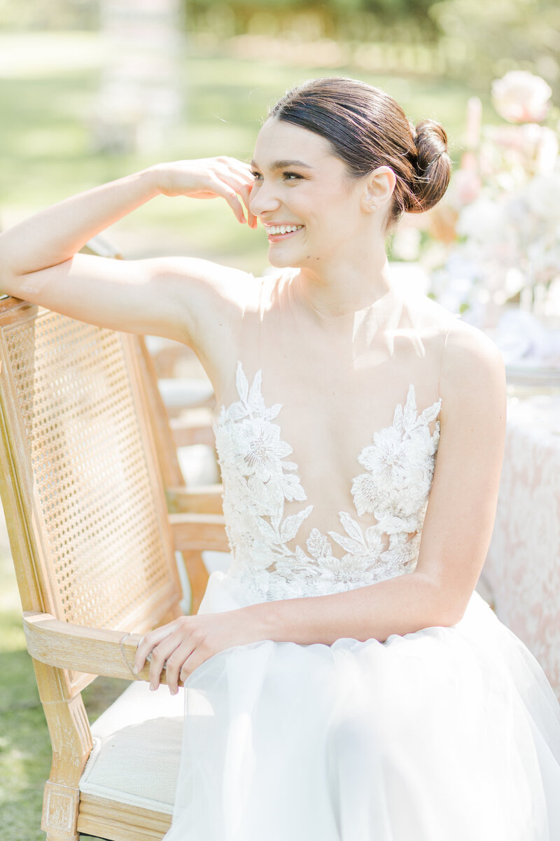 Bride in an illusion wedding dress embellished with lace florals is sitting in a light-tone wood and white upholstered chair for a casual wedding day portrait.. Her arm is resting on the back of the chair and she is smiling off in the distance. Captured by top North Shore Boston and Ipswitch wedding photographer Lia Rose Weddings