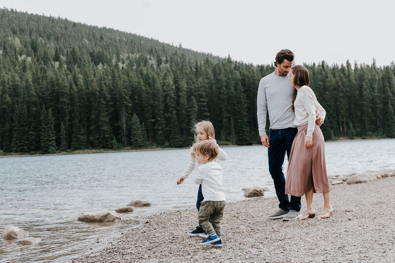 A family enjoys throwing rocks into the river together during their family photography session. The man and woman are holding each other in a loving side hug and the young girl and boy are searching for rocks