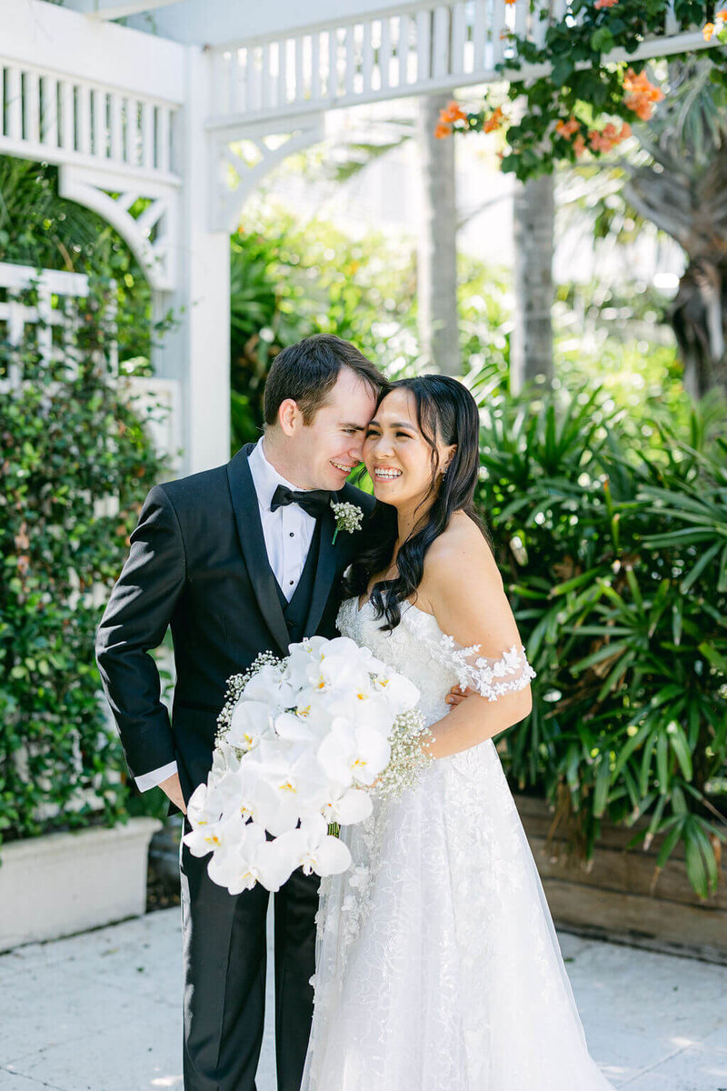 The groom and bride, with her holding a bouquet of white orchids, share genuine laughter during their outdoor wedding photoshoot.