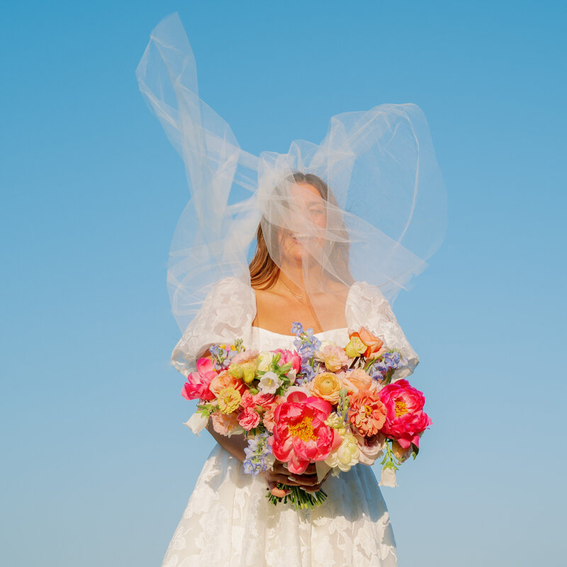 Portrait of a bride holding a big colorful bouquet with her veil flying in her face with a bright blue sky background