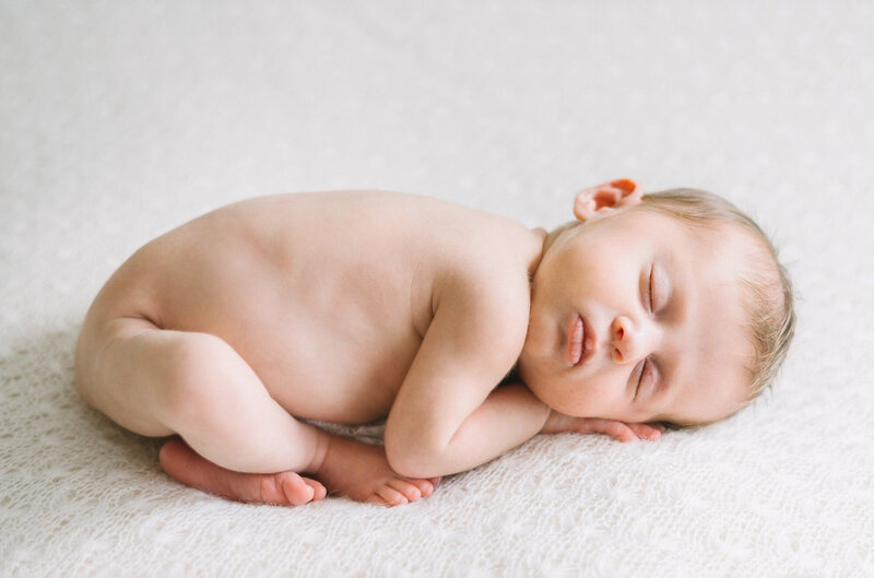 A baby curled up and sleeping on a lace white blanket in pretty natural light.