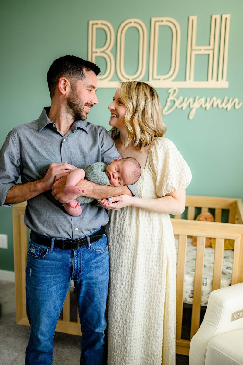 father holds his newborn son while looking at his wife in a nursery