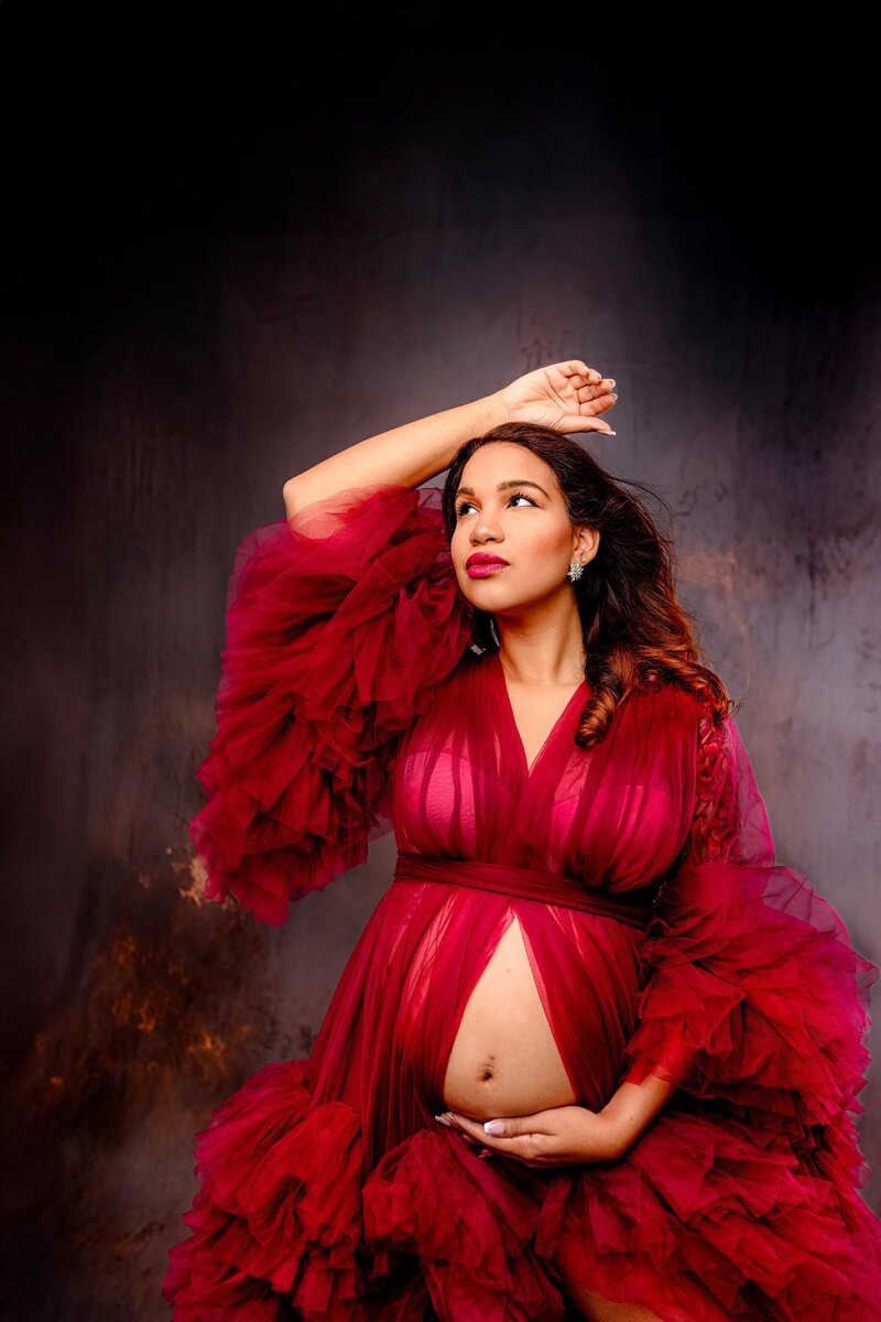 Close up image of a pregnant mom with one arm above her head the other touching her belly looking off camera wearing a red robe