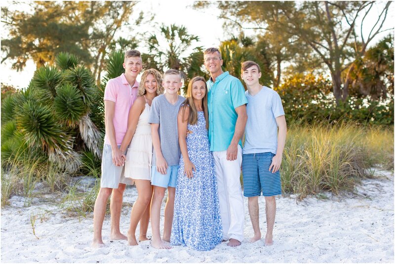 Family standing in white sands smile for a photo with a lush green backdrop.