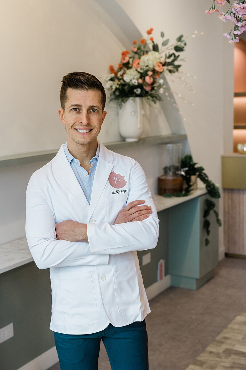Dr. Michael, of Little Chompers, poses in his branded signature white coat, in his Andersonville dentist office. Florals and air plants dot the midcentury modern shelving behind him.