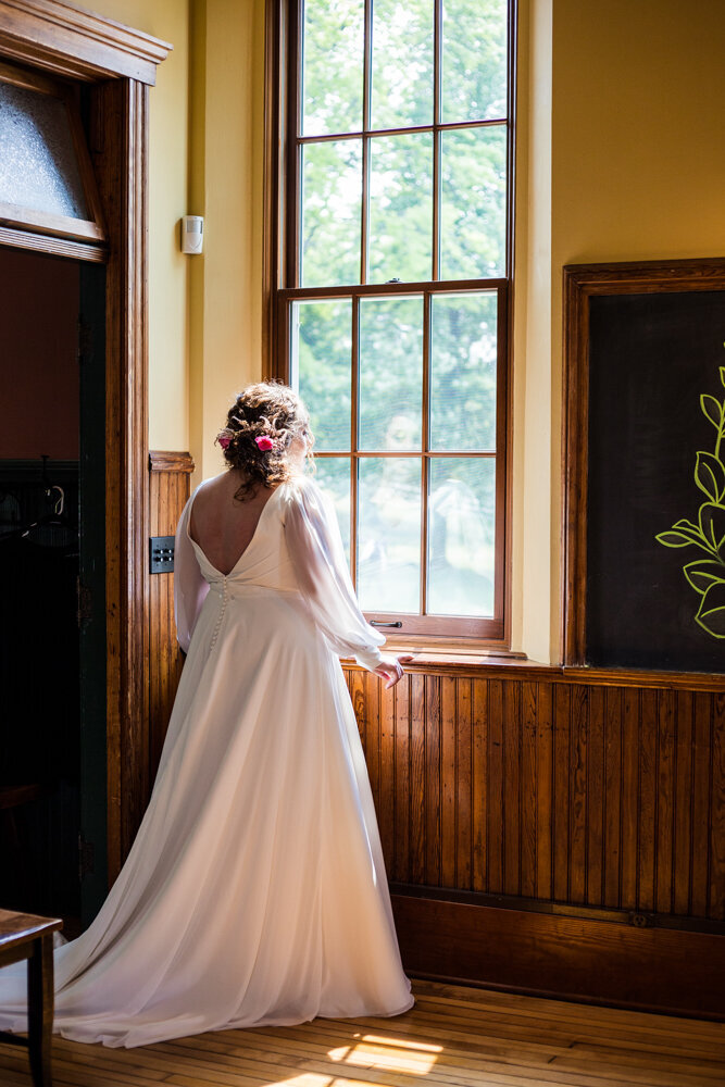 Bride in her dress stands looking out an open window at the Schoolhouse before the start of wedding ceremony