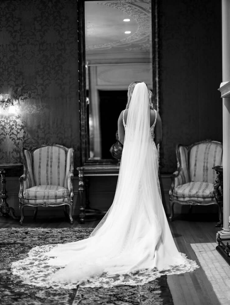 Bride ,wearing long  veil and intricate lace train, looks at herself in a large mirror at Warner Theatre