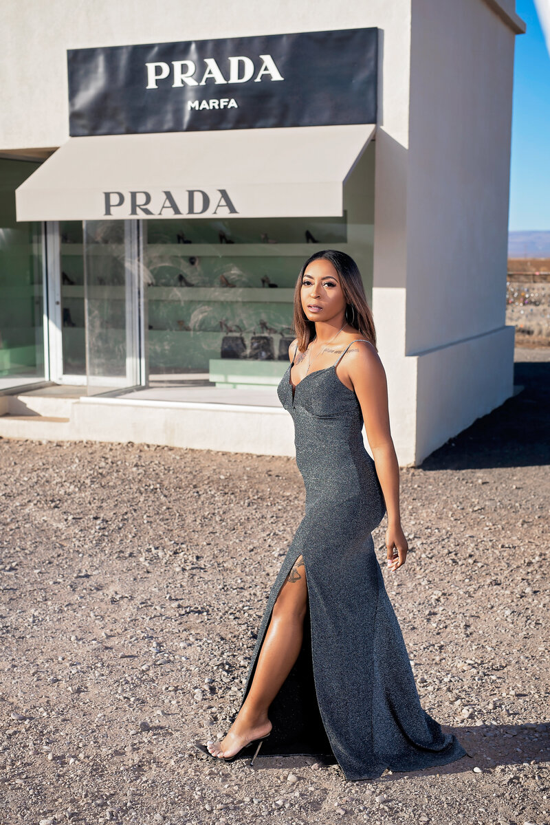 Woman poses in front of the Prada Marfa desert storefront in a dark grey sparkling gown with a leg slit.